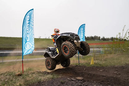 Person riding a black CFMOTO ATV in mid-air on a dirt track with two blue CFMOTO flags in the background.