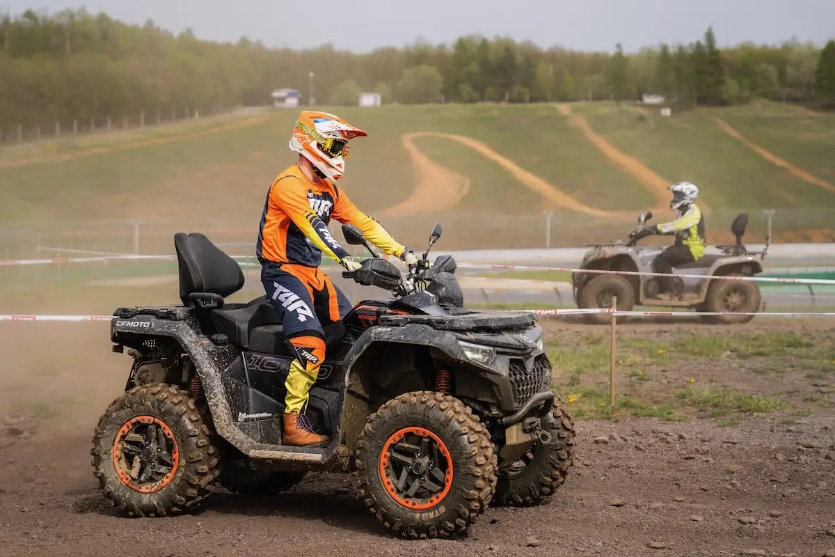 Two people on CFMOTO ATVs on a dirt race track with trees in the background