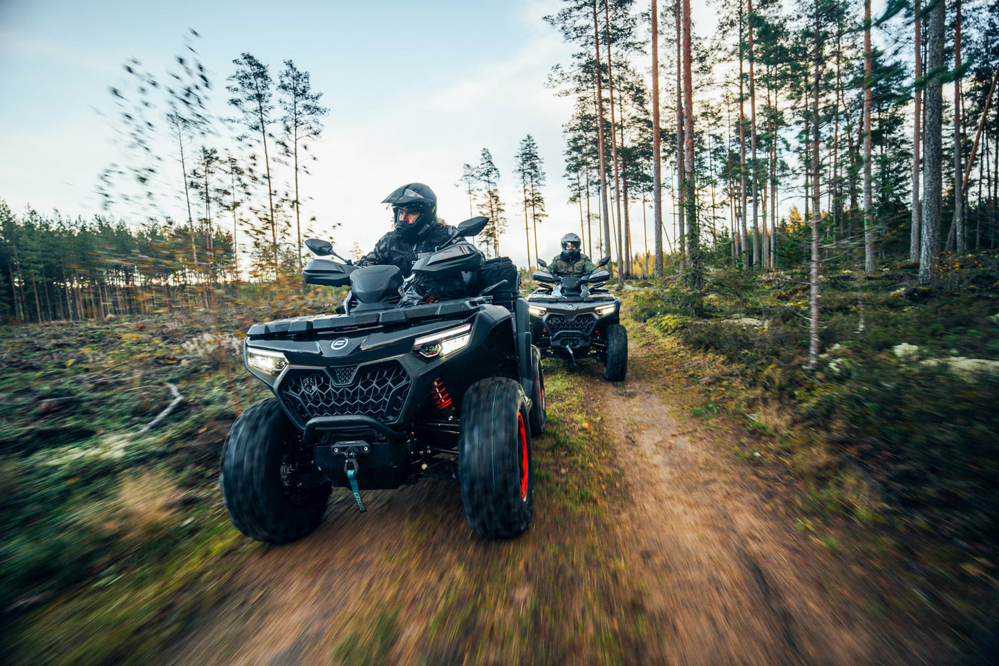 Two people riding CFMOTO ATVs on a dirt path through a forest.