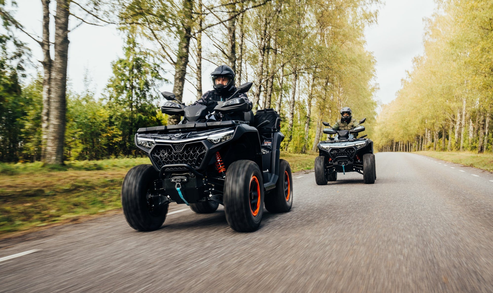 Two people riding CFMOTO ATVs on a forest road with trees lining the sides.