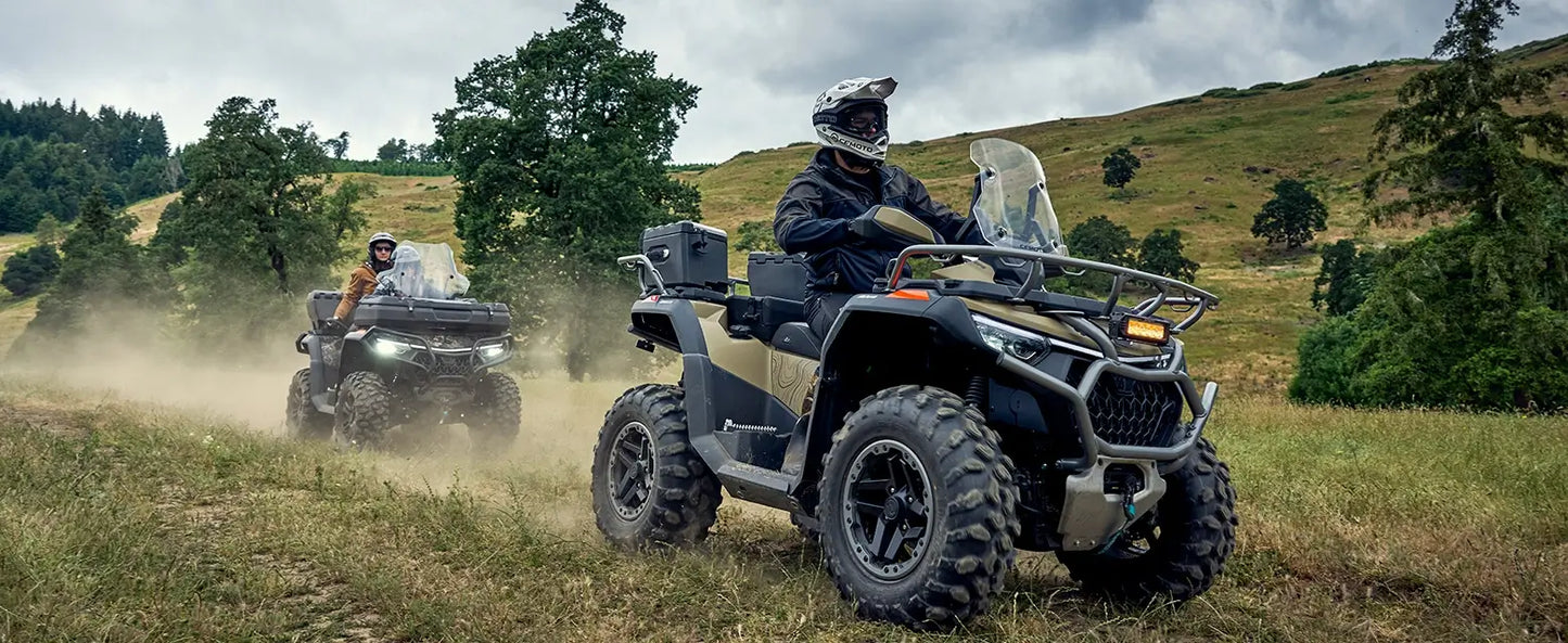Two CFMOTO off-road vehicles driving through a grassy field with trees in the background.