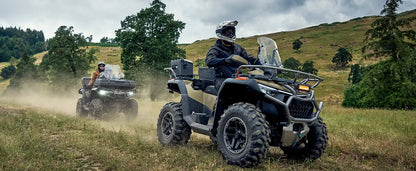 Two CFMOTO off-road vehicles driving through a grassy field with trees in the background.