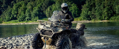 Person riding a CFMOTO ATV through a river with a forested area in the background