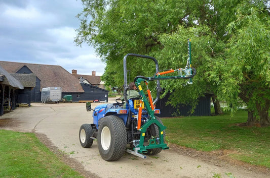 Small tractor with a large Wessex hedge cutter attachment on a dirt road next to a building and trees.