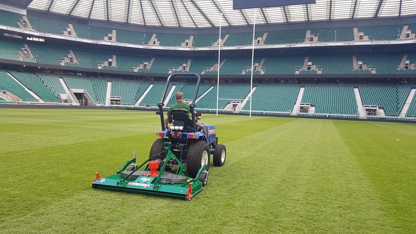 Person operating a wessex mower on a large green field with stadium seats in the background