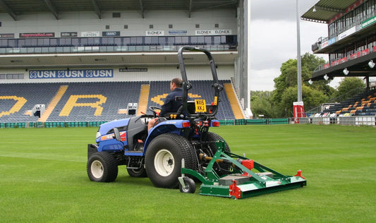 Person operating a wessex mower on a sports field with stadium seating in the background.