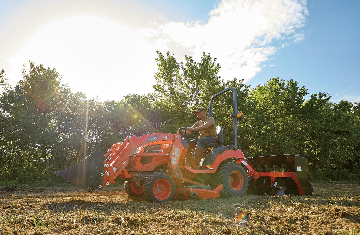 Person operating a Kioti tractor in a field with trees in the background