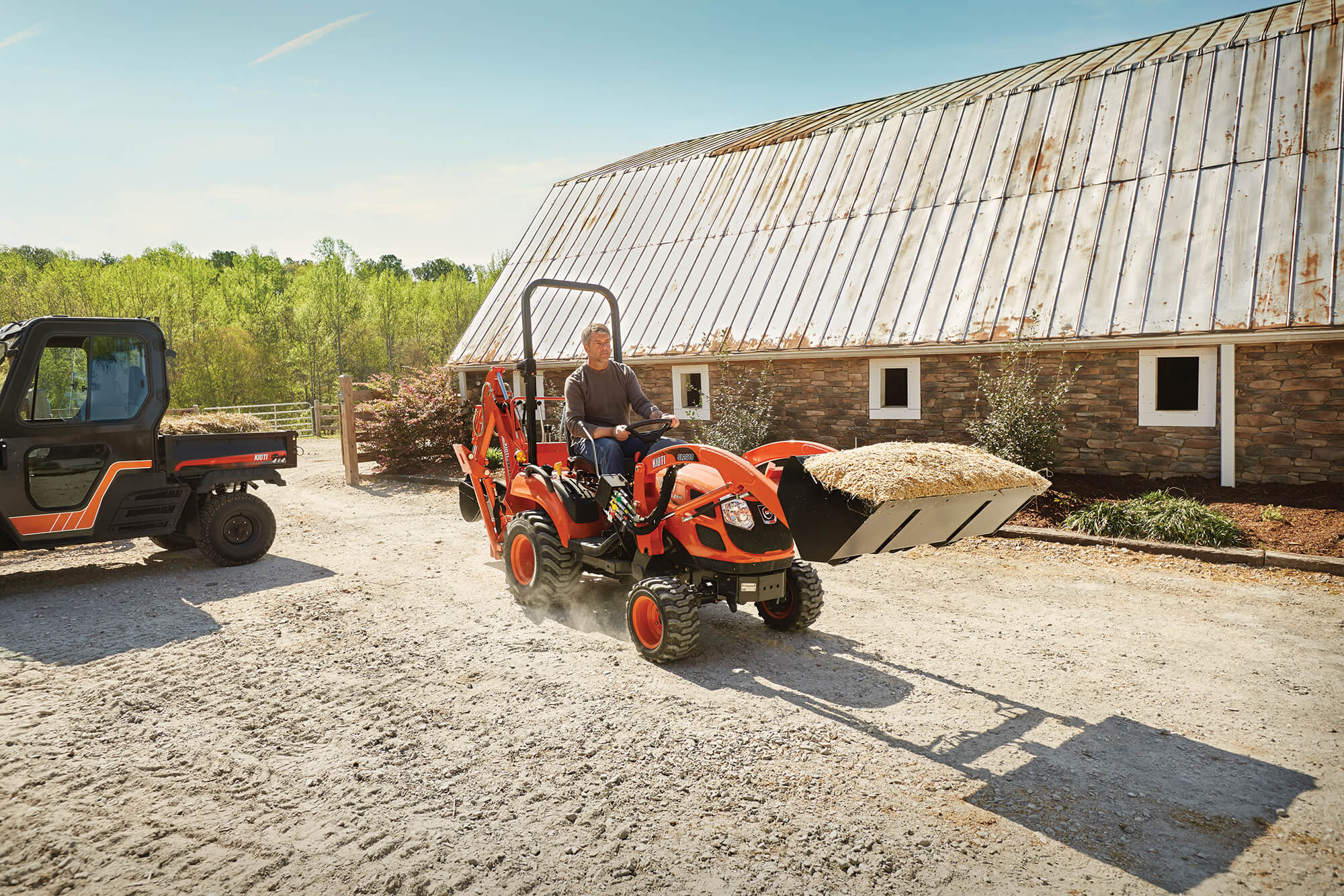 Person operating a Kioti compact tractor in front of a barn.