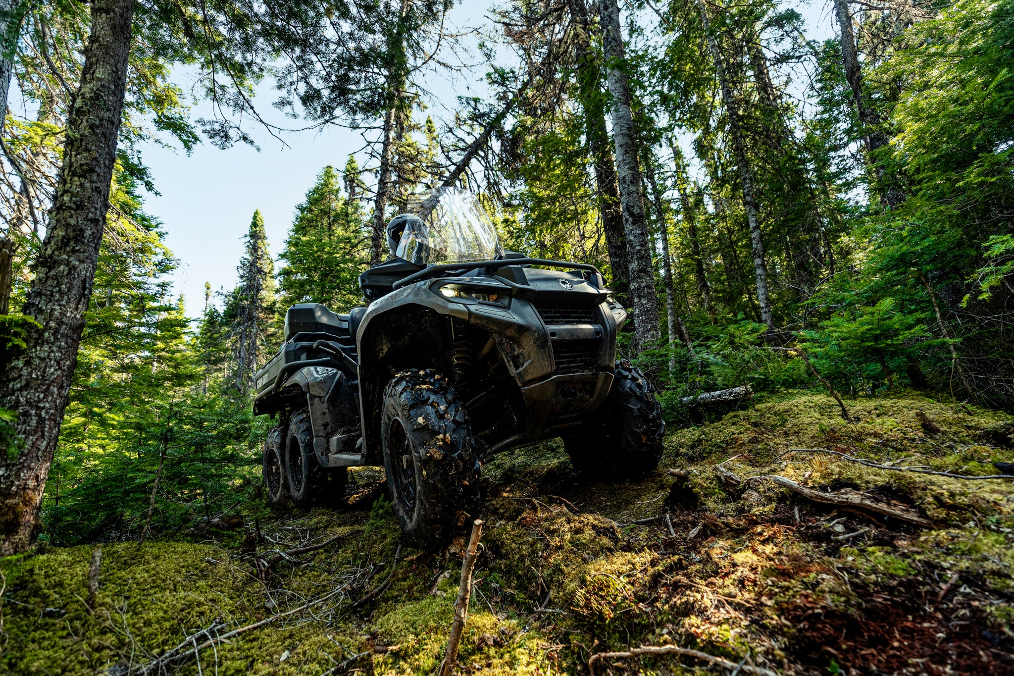 Can-Am Outlander 6x6 ATV driving through a forest with trees and greenery around