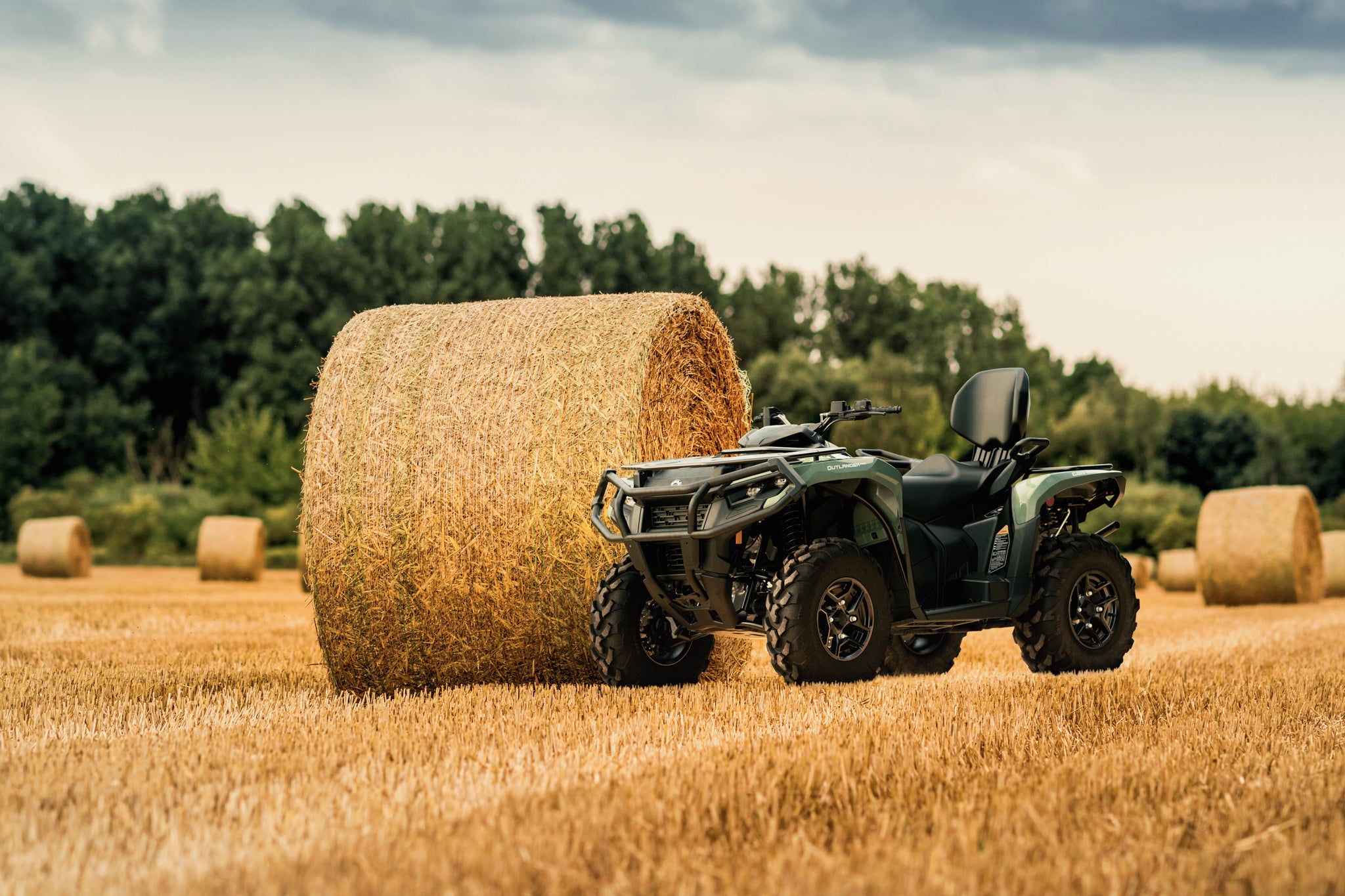 Can-Am Outlander ATV parked next to a hay bale in a field with trees in the background