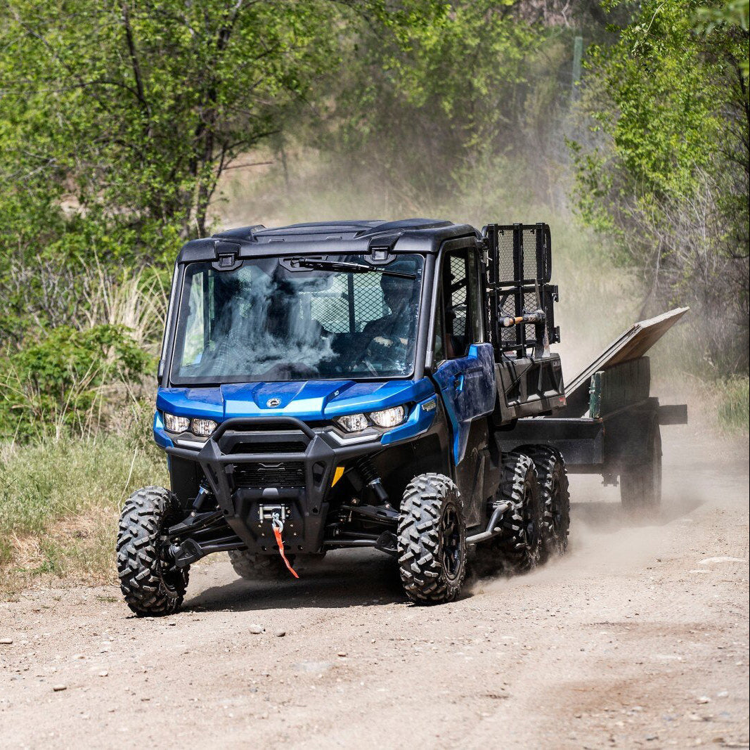 Blue Can-Am Traxter 6x6 UTV with a flatbed trailer on a dirt road surrounded by greenery