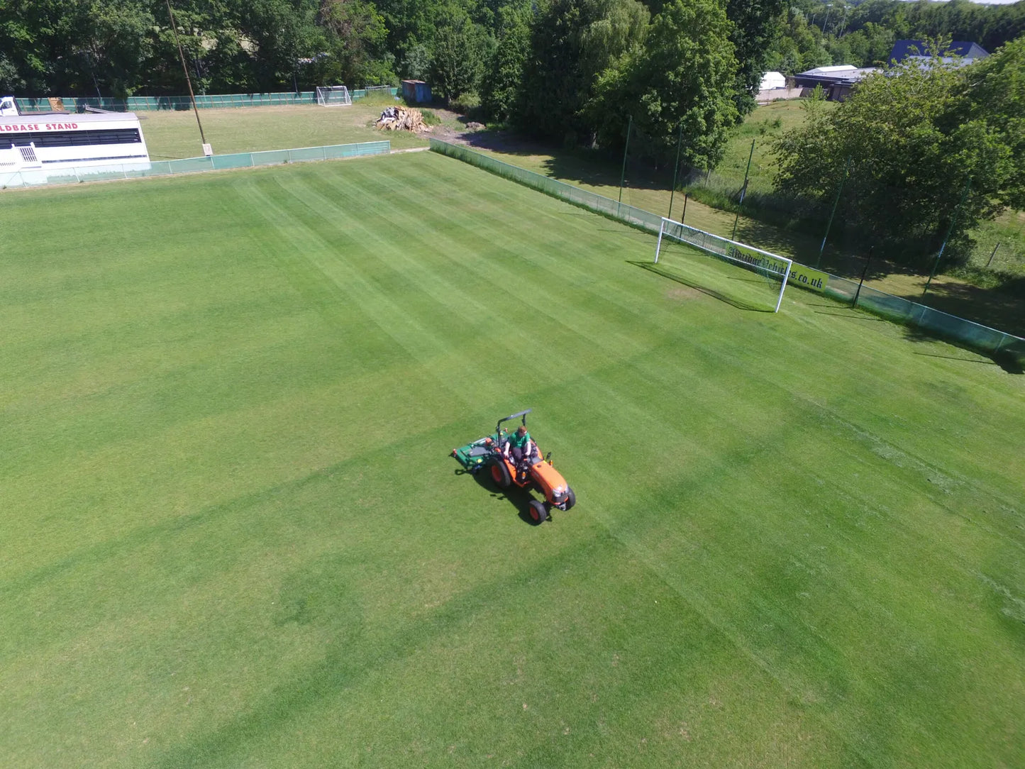 Person operating a wessex mower on a well-maintained soccer field with trees and buildings in the background.