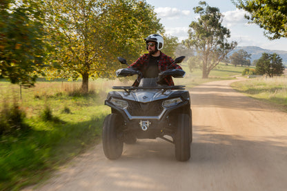 Person riding a CFMOTO quad bike on a dirt road with trees and mountains in the background