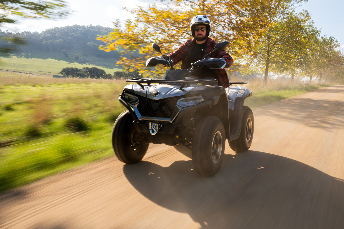 Person riding a CFMOTO ATV on a dirt road with trees and fields in the background