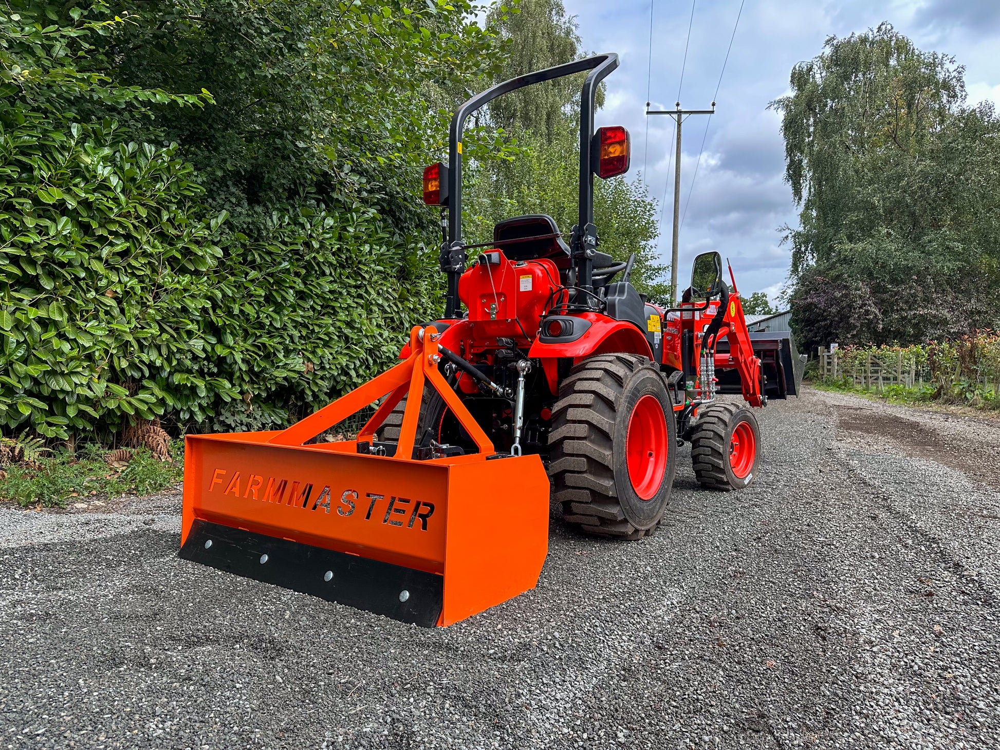 FarmMaster tractor box grader attachment on a gravel road surrounded by trees.