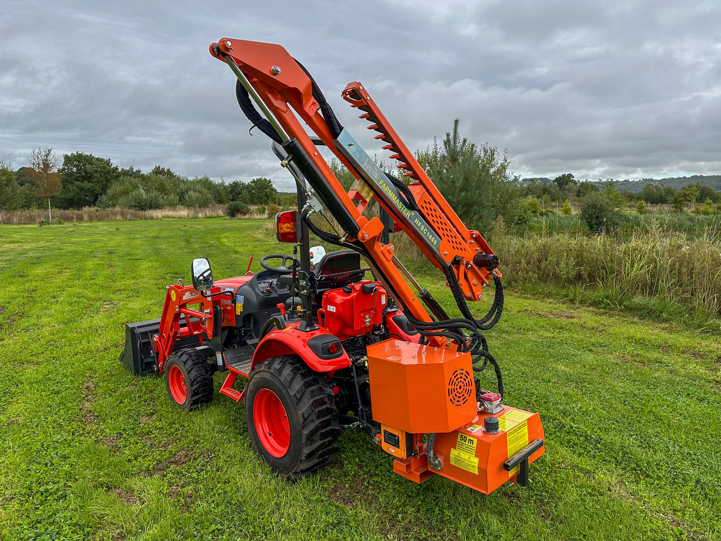 Kioti compact tractor with a large FarmMaster tractor hedge cutter attachment in a grassy field on a cloudy day