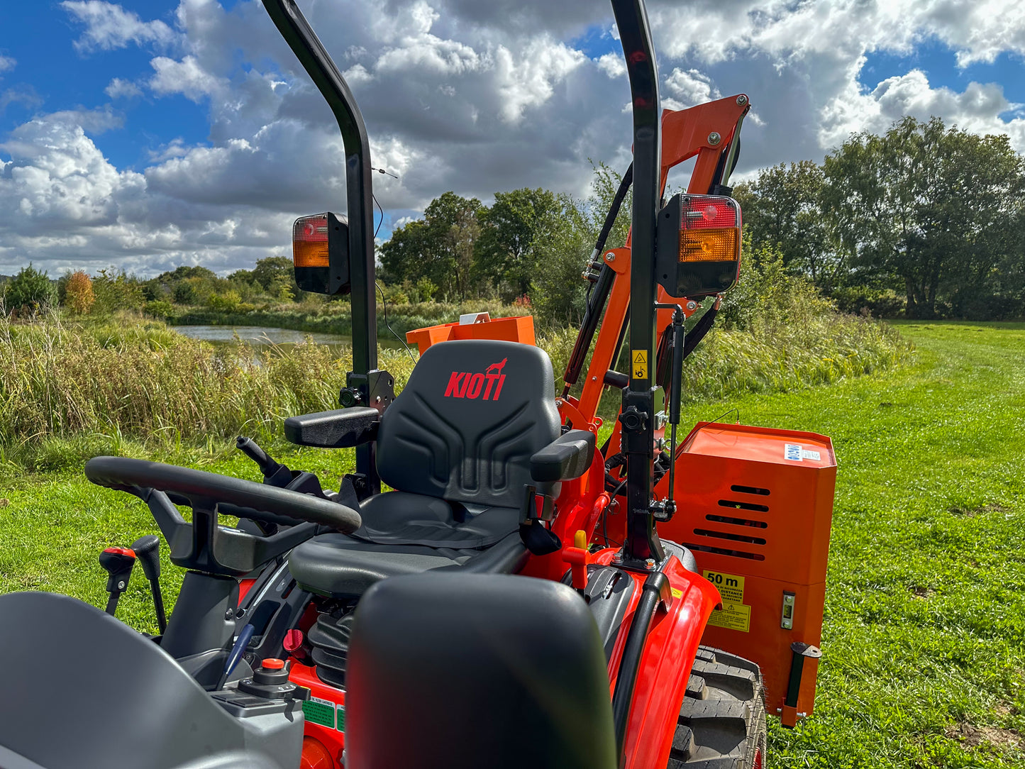 Kioti tractor and FarmMaster tractor hedge cutter in a grassy field with trees and blue sky.