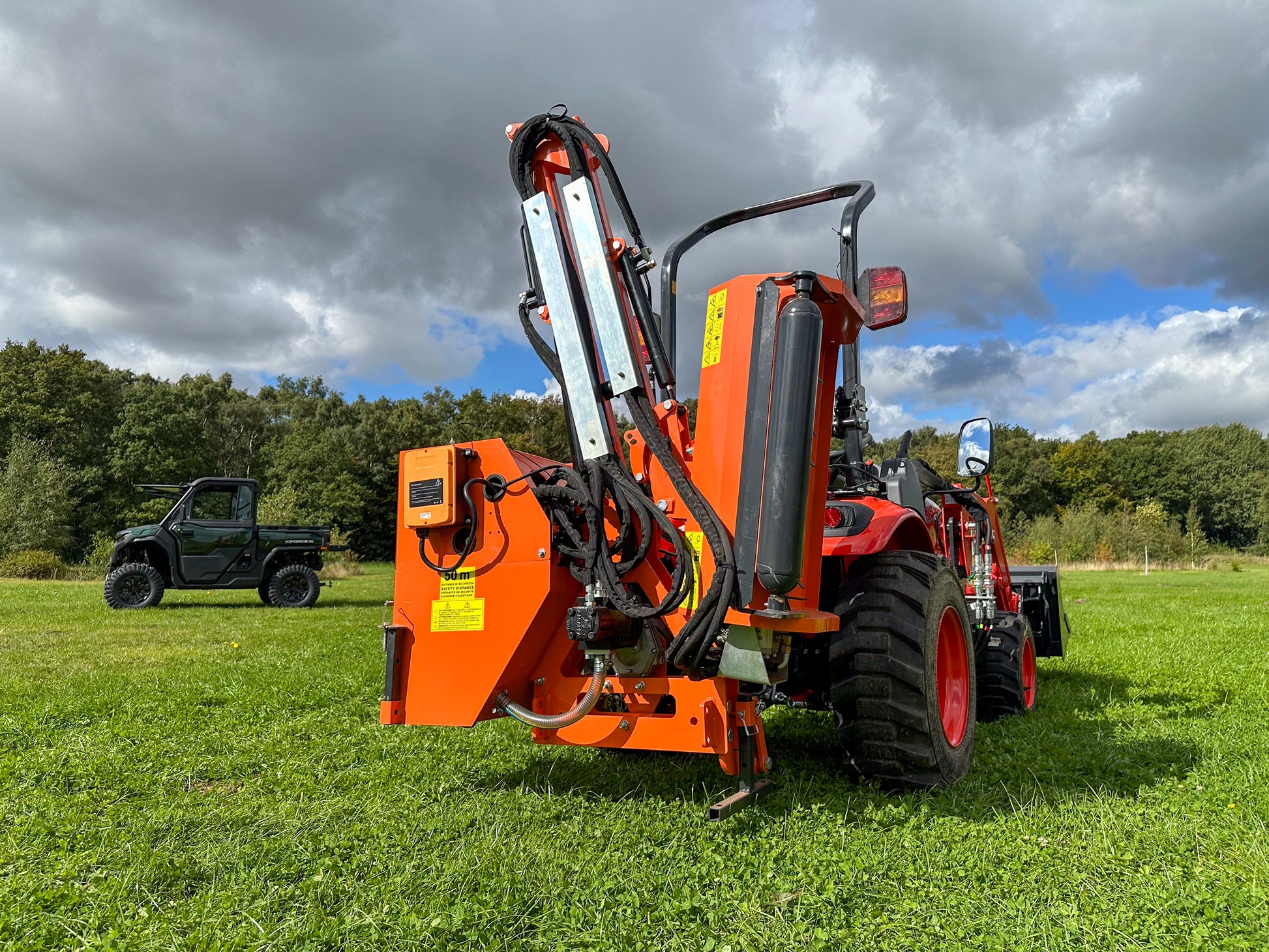 Kioti tractor with a FarmMaster tractor hedge cutter on a grassy field under a cloudy sky.