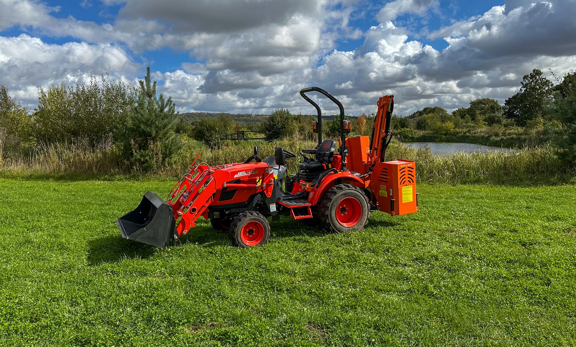 FarmMaster tractor hedge cutter on a grassy field with trees and clouds in the background