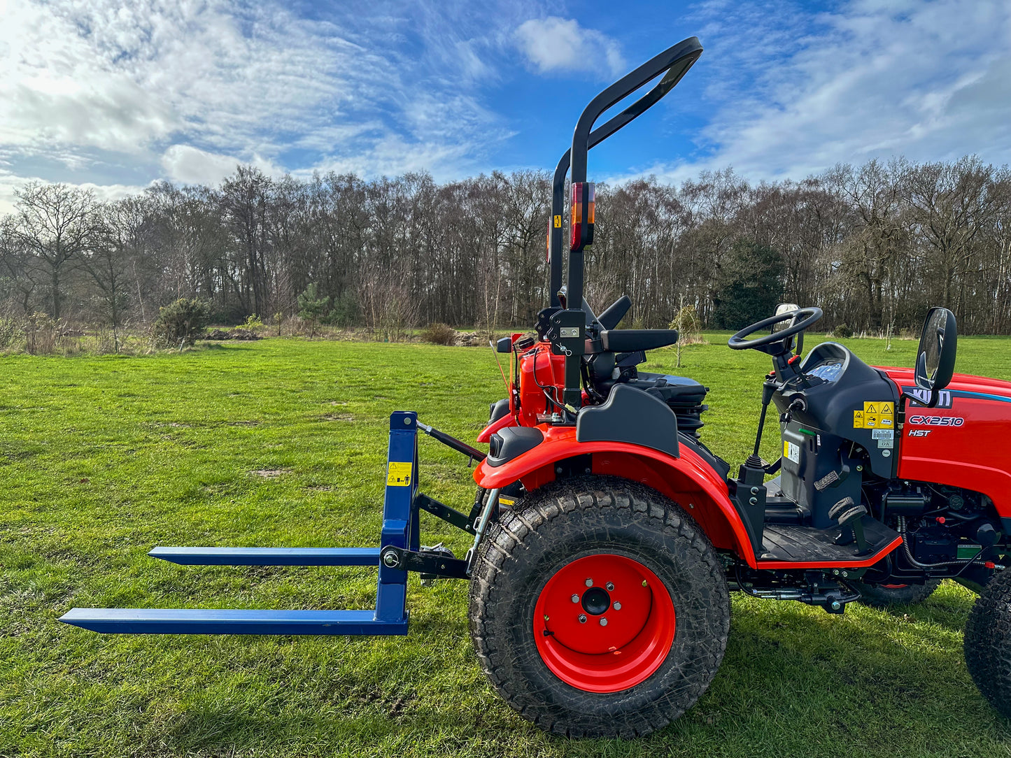 Kioti tractor with a FarmMaster tractor pallet forks on a grassy field with trees in the background.