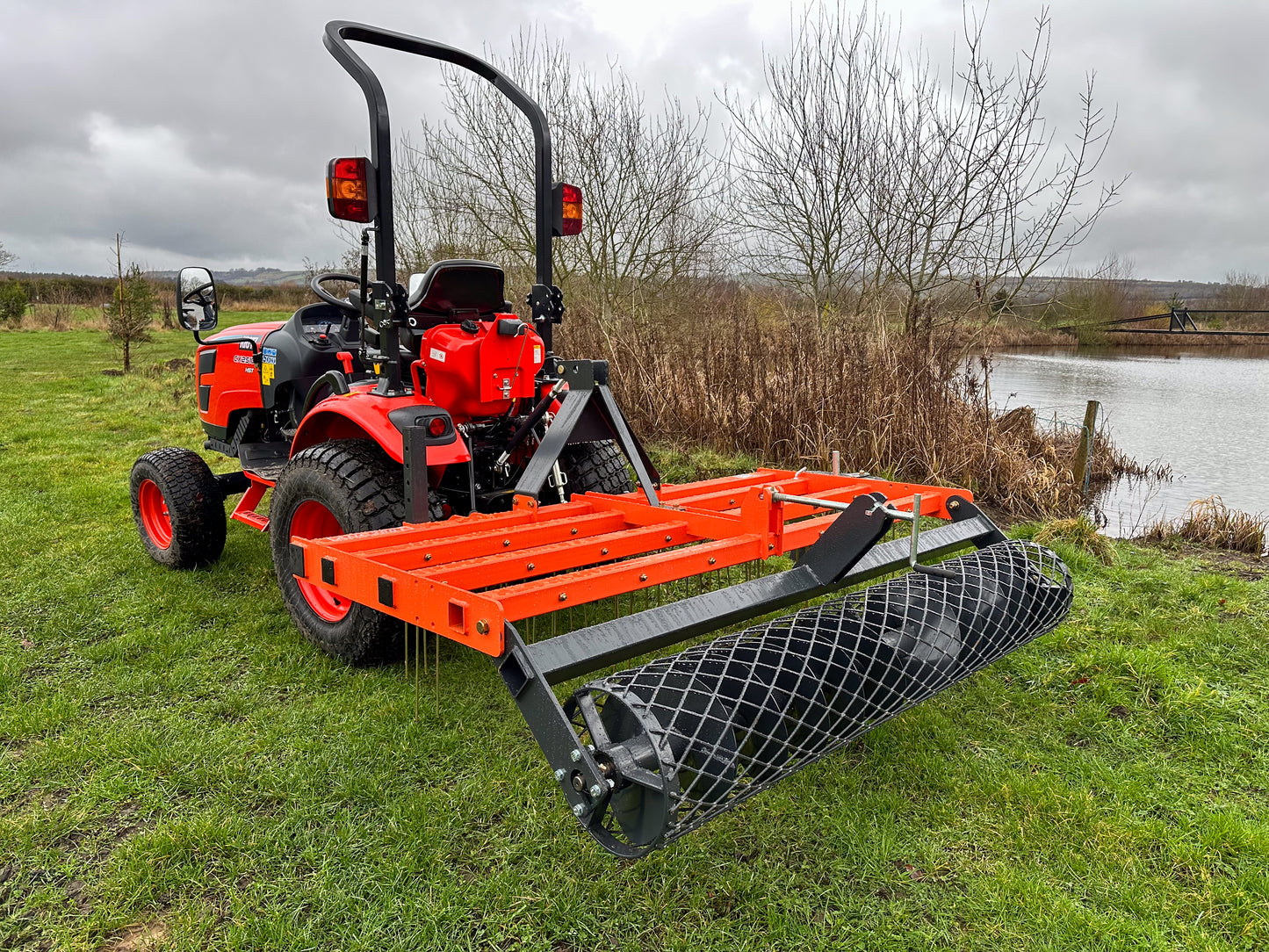 Kioti compact tractor with a FarmMaster tractor arena conditioner on a grassy field near a body of water.
