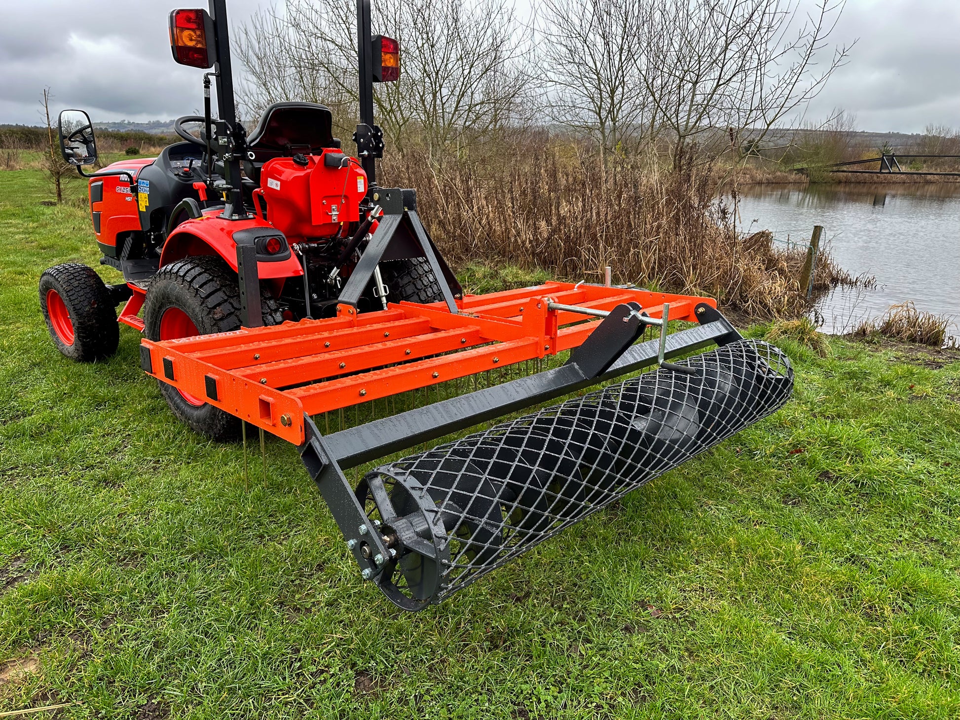 Kioti compact tractor with a FarmMaster tractor arena conditioner on a grassy field near a body of water.