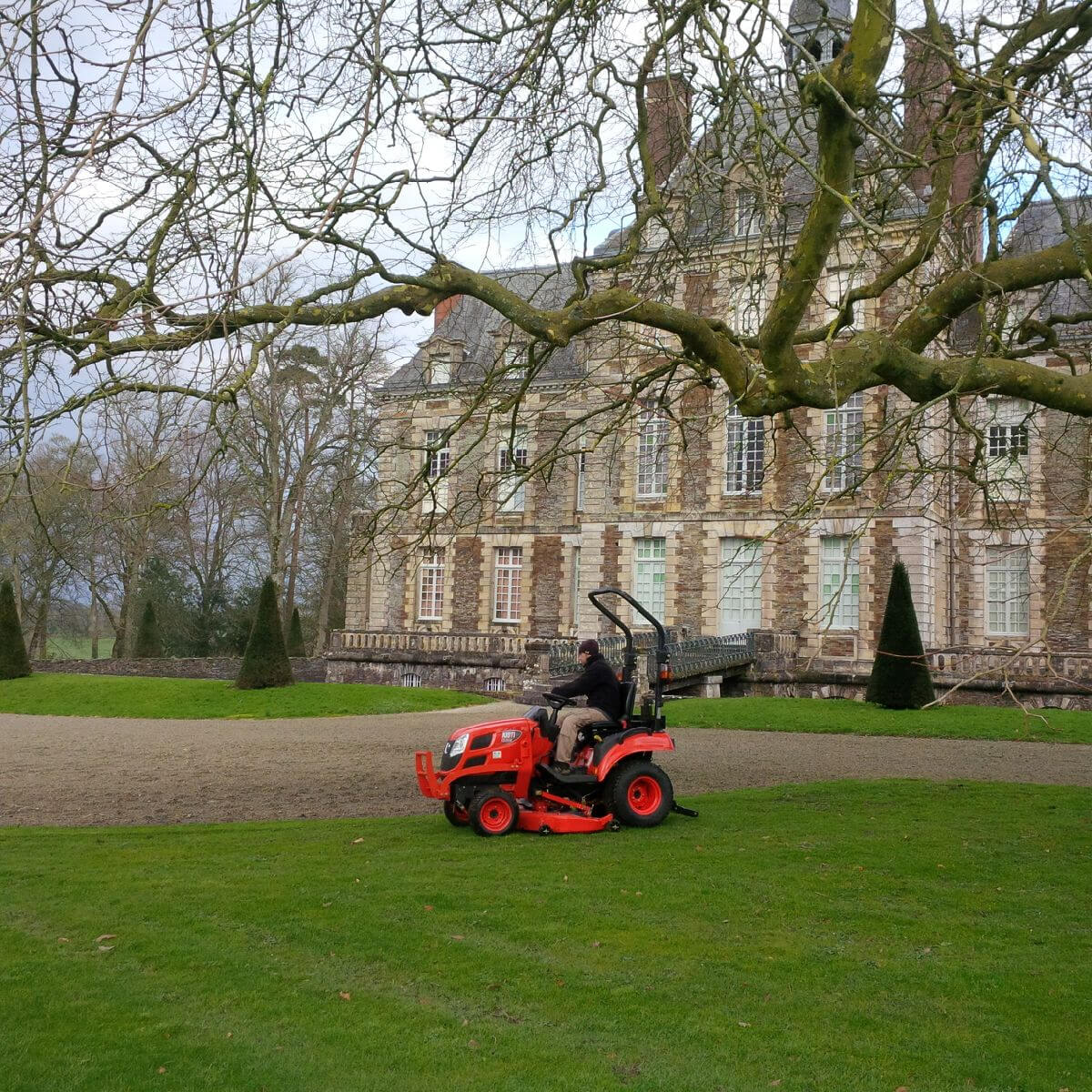 Person operating a Kioti sub-compact tractor in front of a large stone building with trees in the foreground.