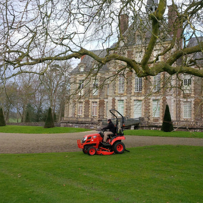 Person operating a Kioti sub-compact tractor in front of a large stone building with trees in the foreground.