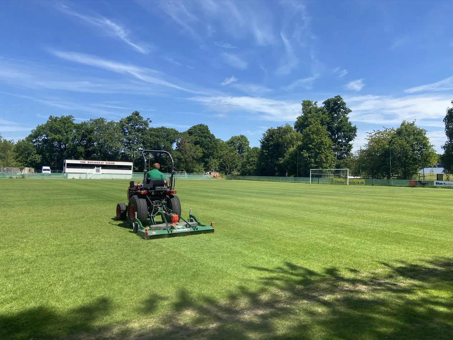 Person operating a Wessex finishing mower on a grass field with trees and buildings in the background.