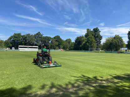 Person operating a Wessex finishing mower on a grass field with trees and buildings in the background.