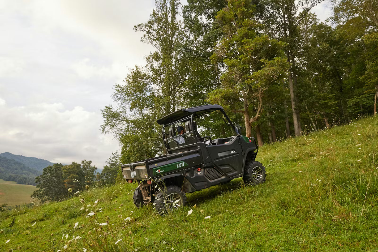 Kioti K9 UTV on a grassy hillside with trees and mountains in the background