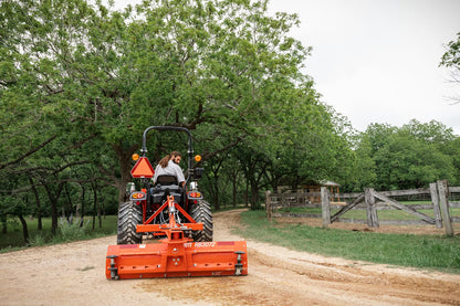 Person operating a Kioti tractor with an attachment on a dirt road in a rural setting.
