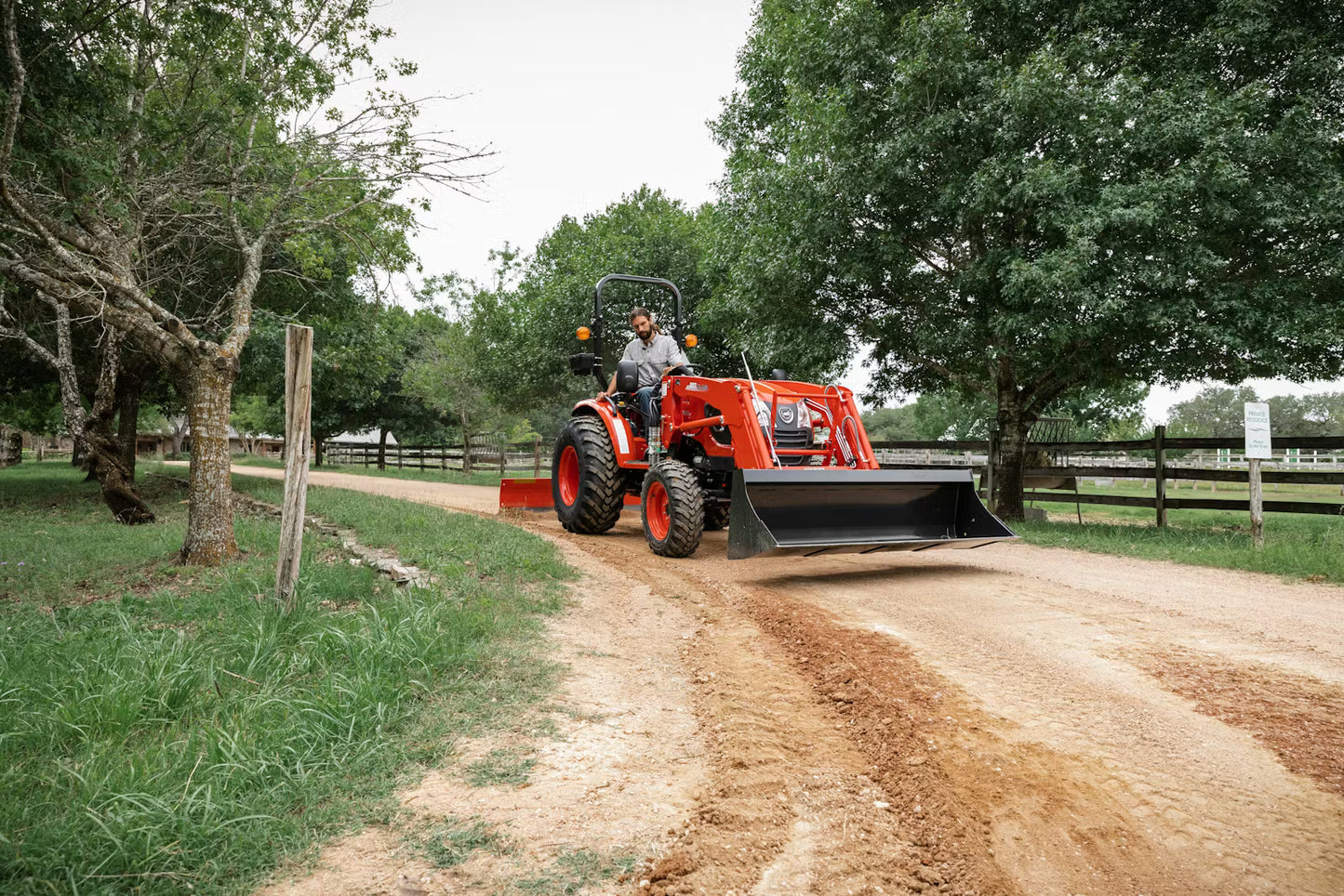 Person operating a red Kioti tractor on a dirt path with trees and a wooden fence in the background
