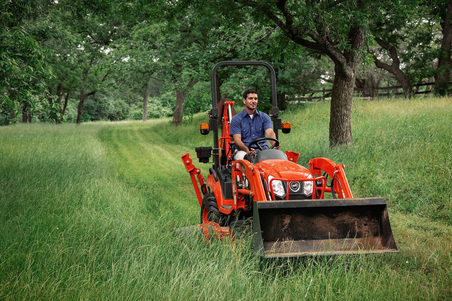 Man operating a Kioti compact tractor with mid-deck mower in a grassy field with trees in the background