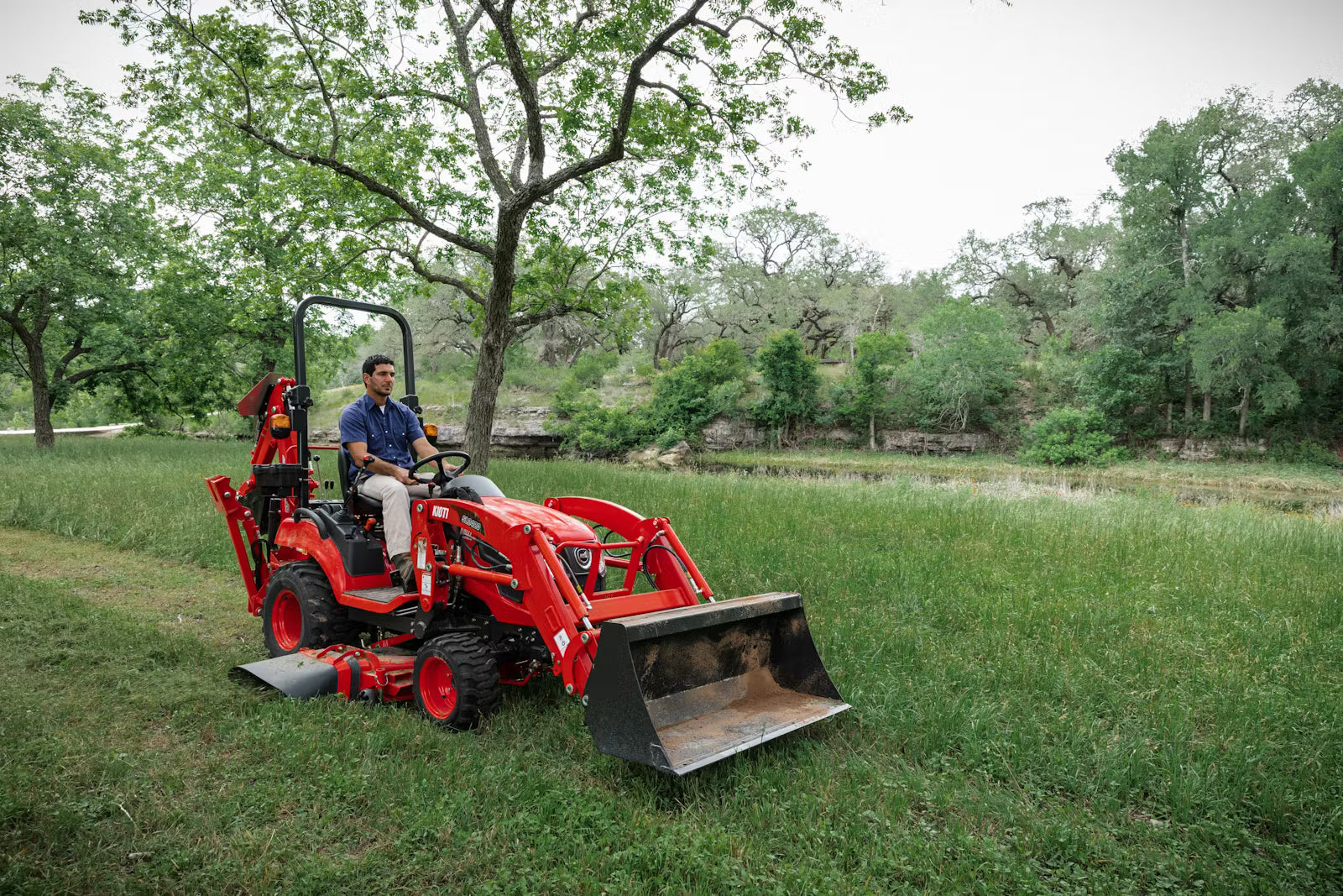 Person operating a Kioti tractor with mower in a grassy field with trees.