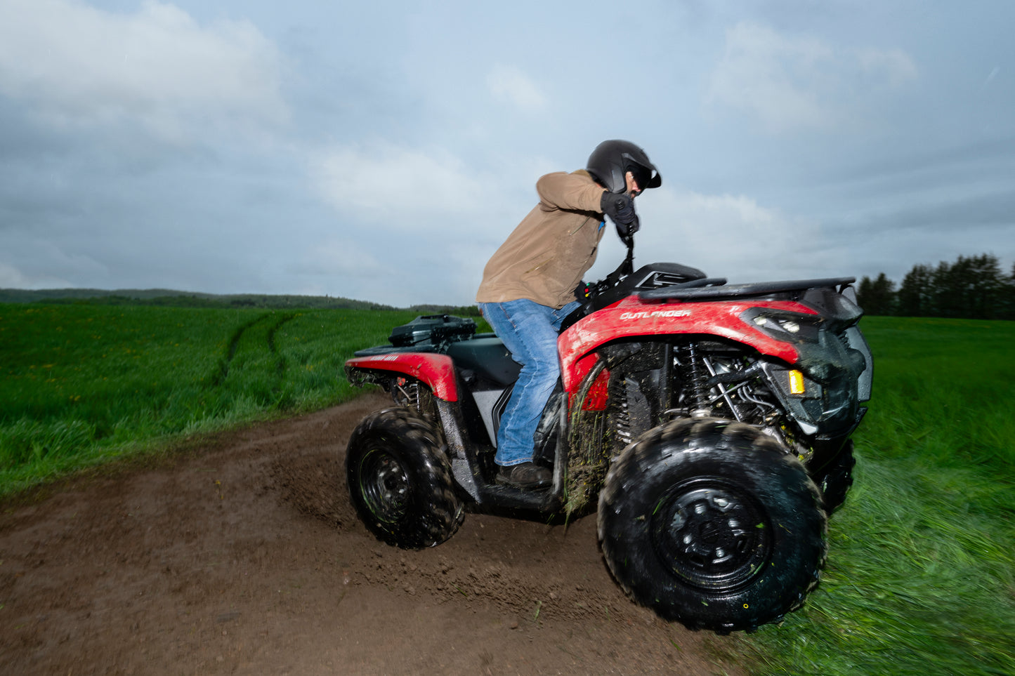 Person riding a red Can-Am ATV on a dirt path with grassy fields and trees in the background.