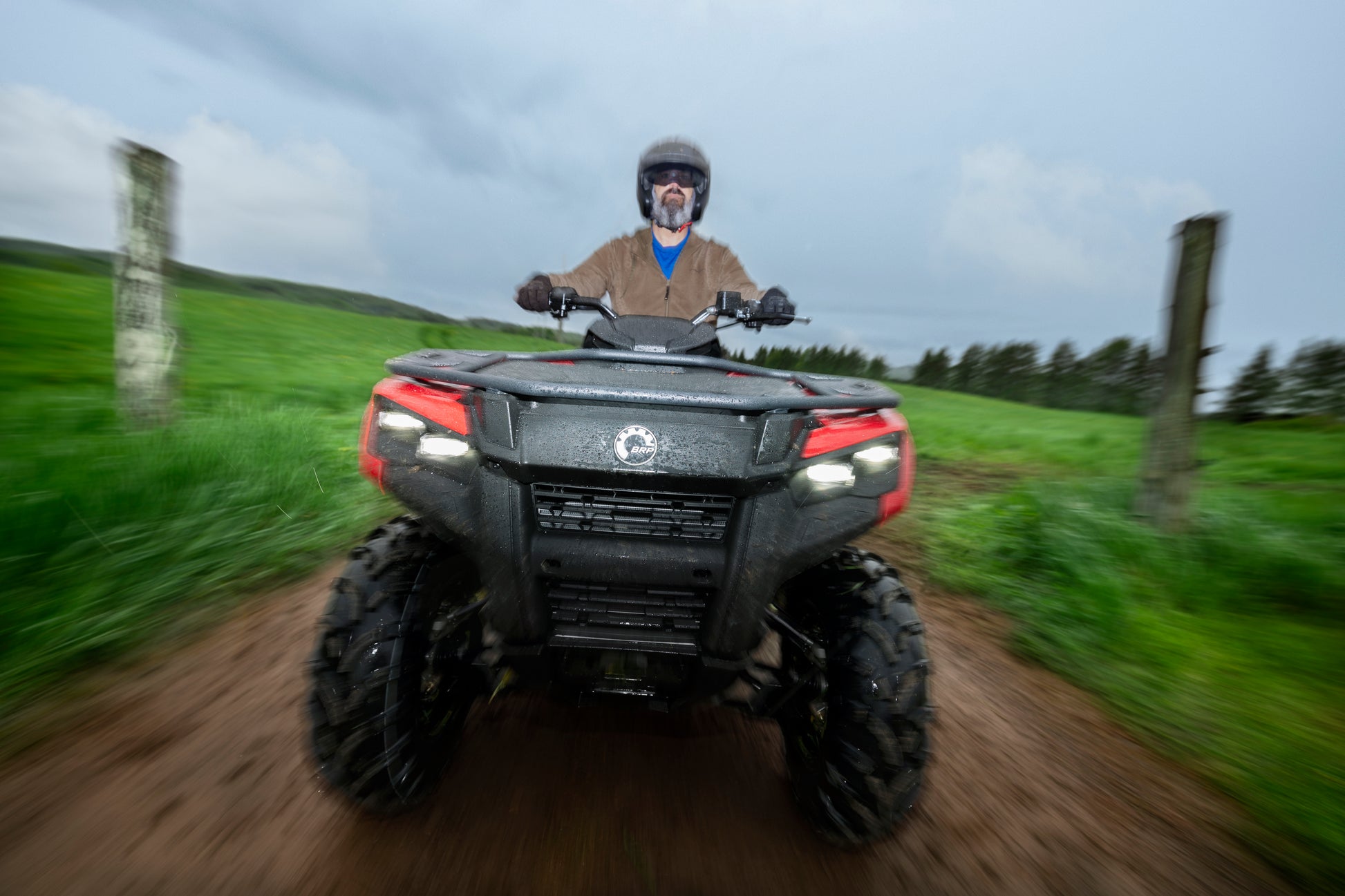 Person riding a Can-Am ATV on a dirt path with green fields and trees in the background