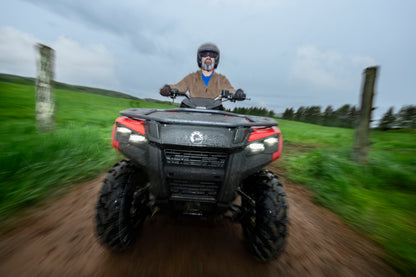 Person riding a Can-Am ATV on a dirt path with green fields and trees in the background