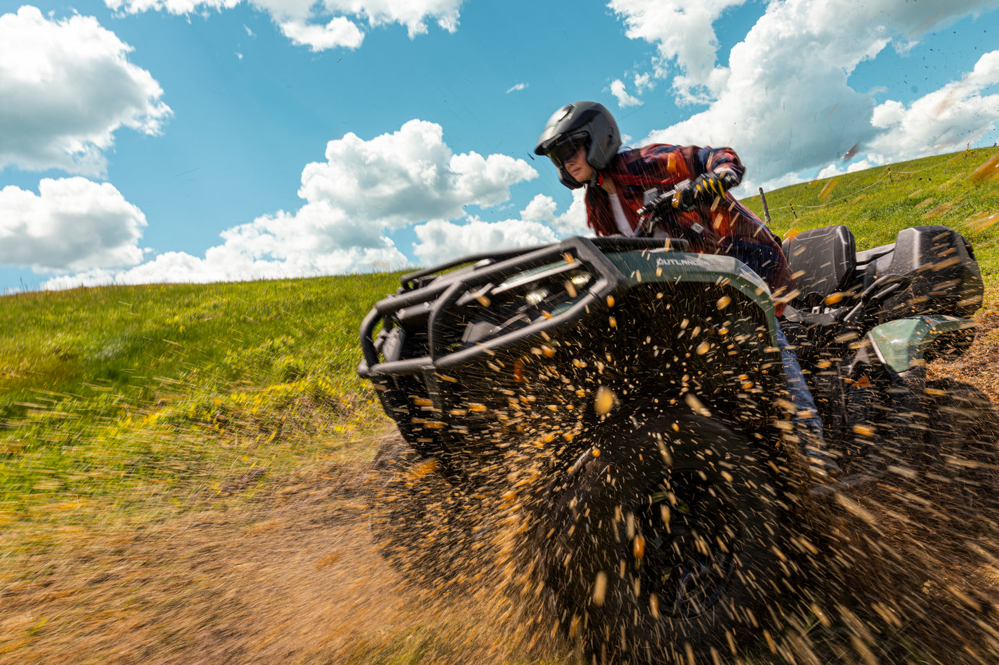 Person riding a Can-Am ATV on a dirt trail with a grassy field and blue sky in the background