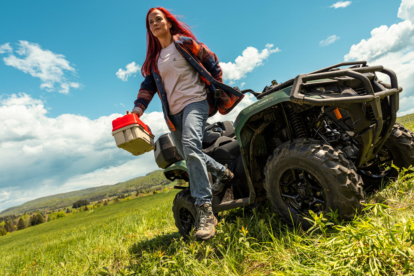 Person with red hair standing on a Can-Am ATV in a grassy field with a blue sky.
