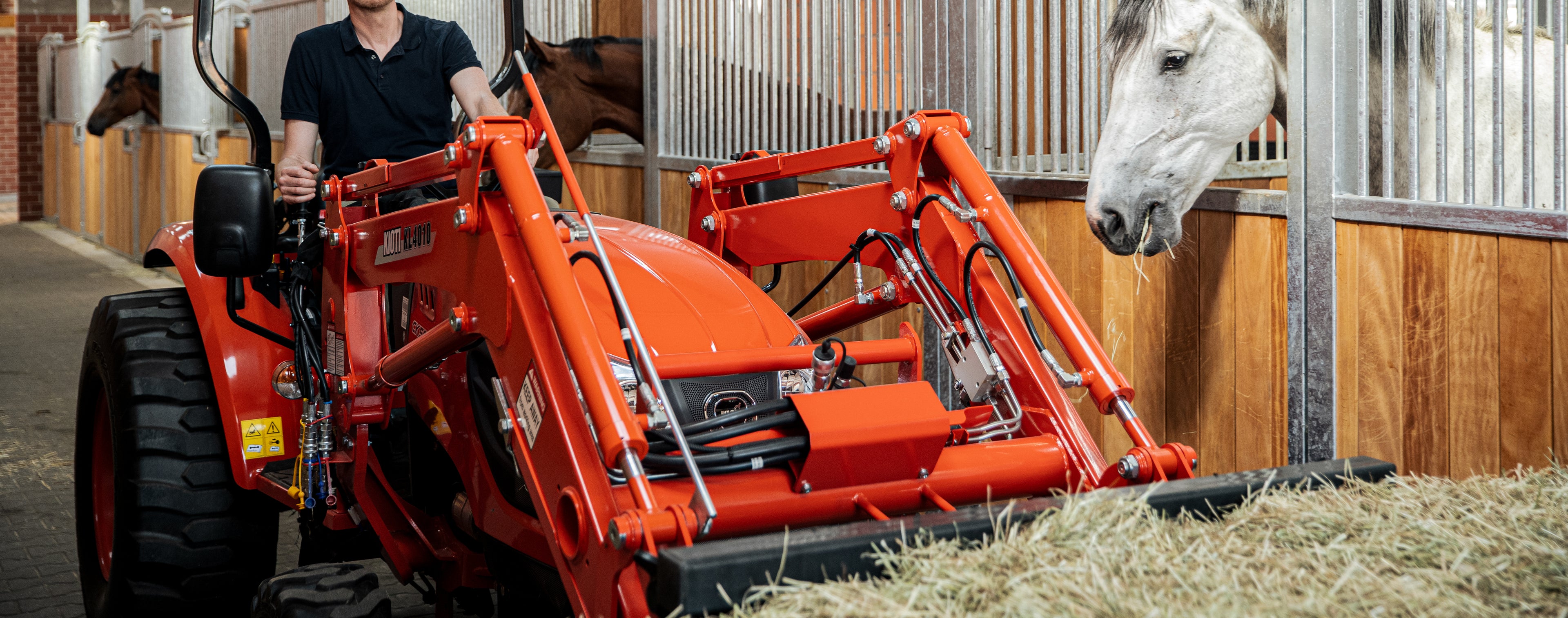 Person operating a Kioti tractor in a stable with horses.