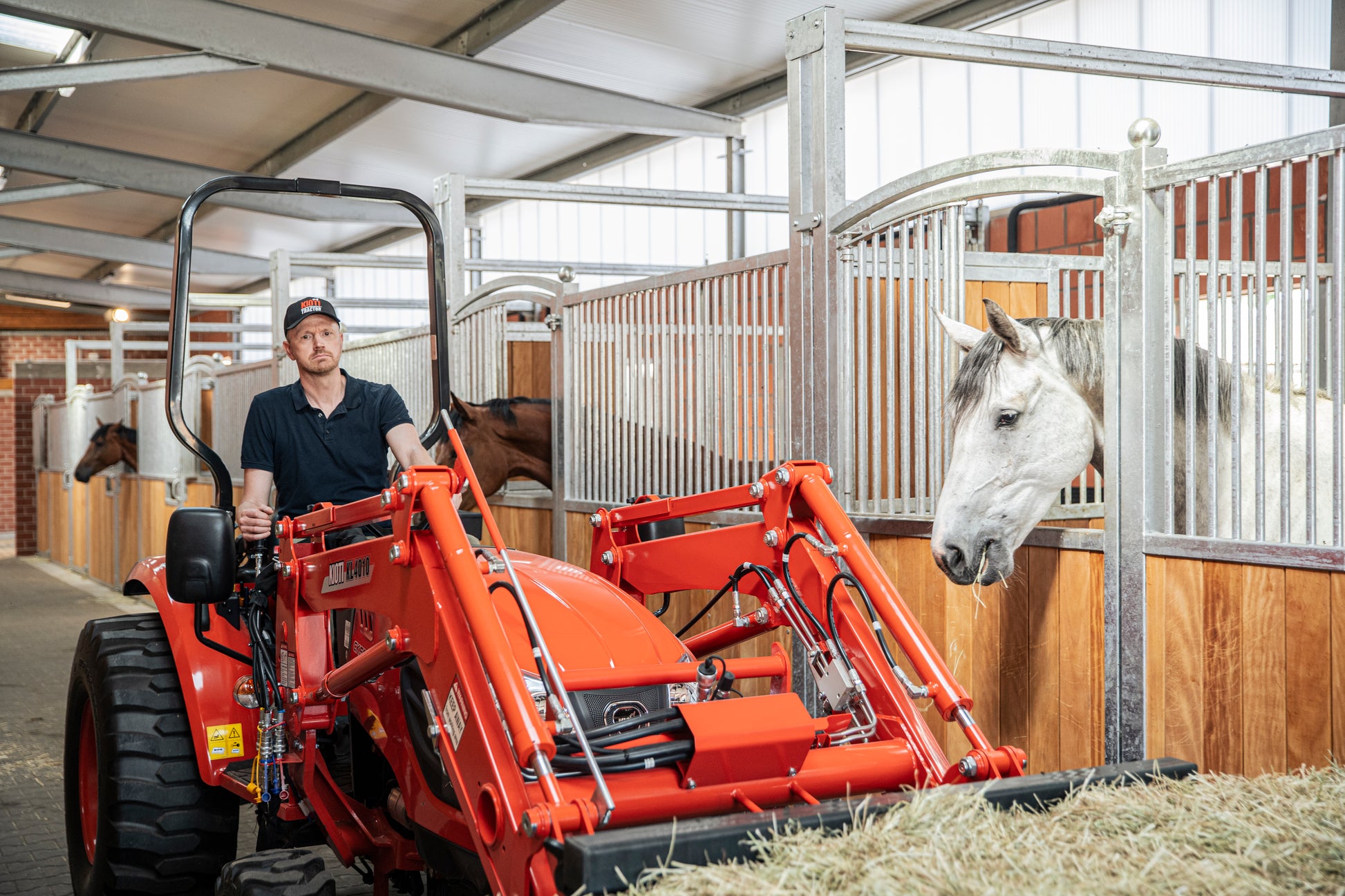 Person operating a red Kioti tractor in a stable with horses.