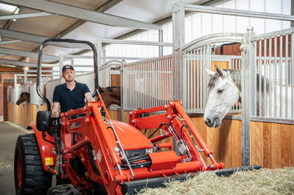 Person operating a red Kioti tractor in a stable with horses.