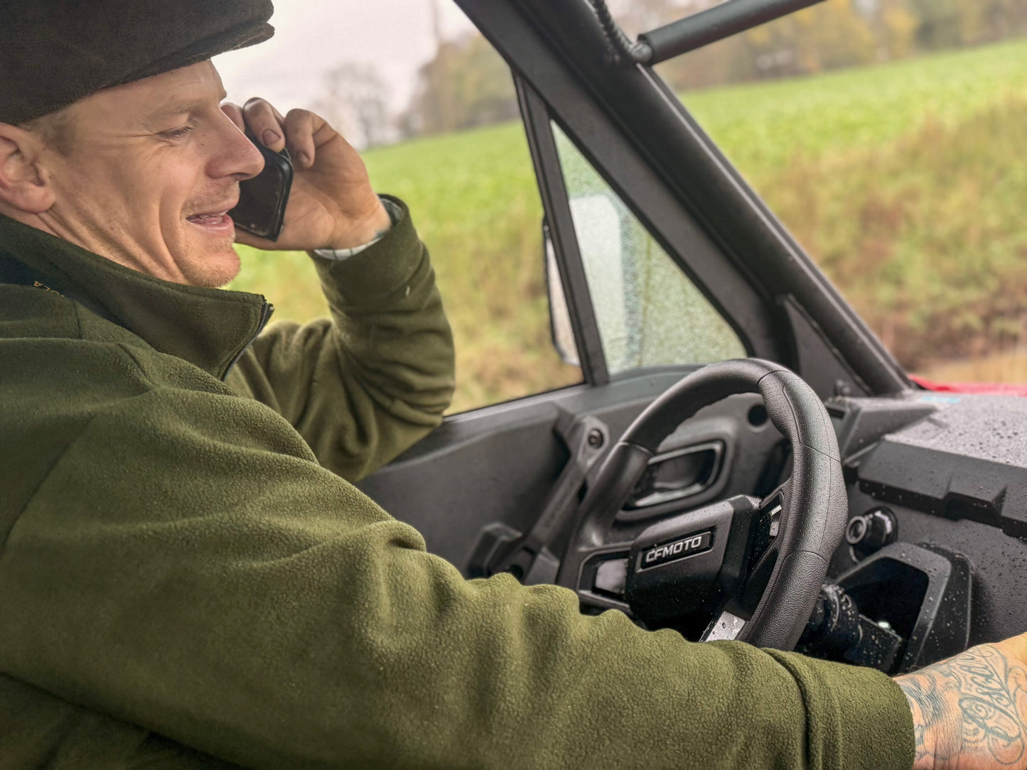 Man in a green jacket sitting inside a CFMOTO off-road vehicle, talking on a phone with a field in the background.