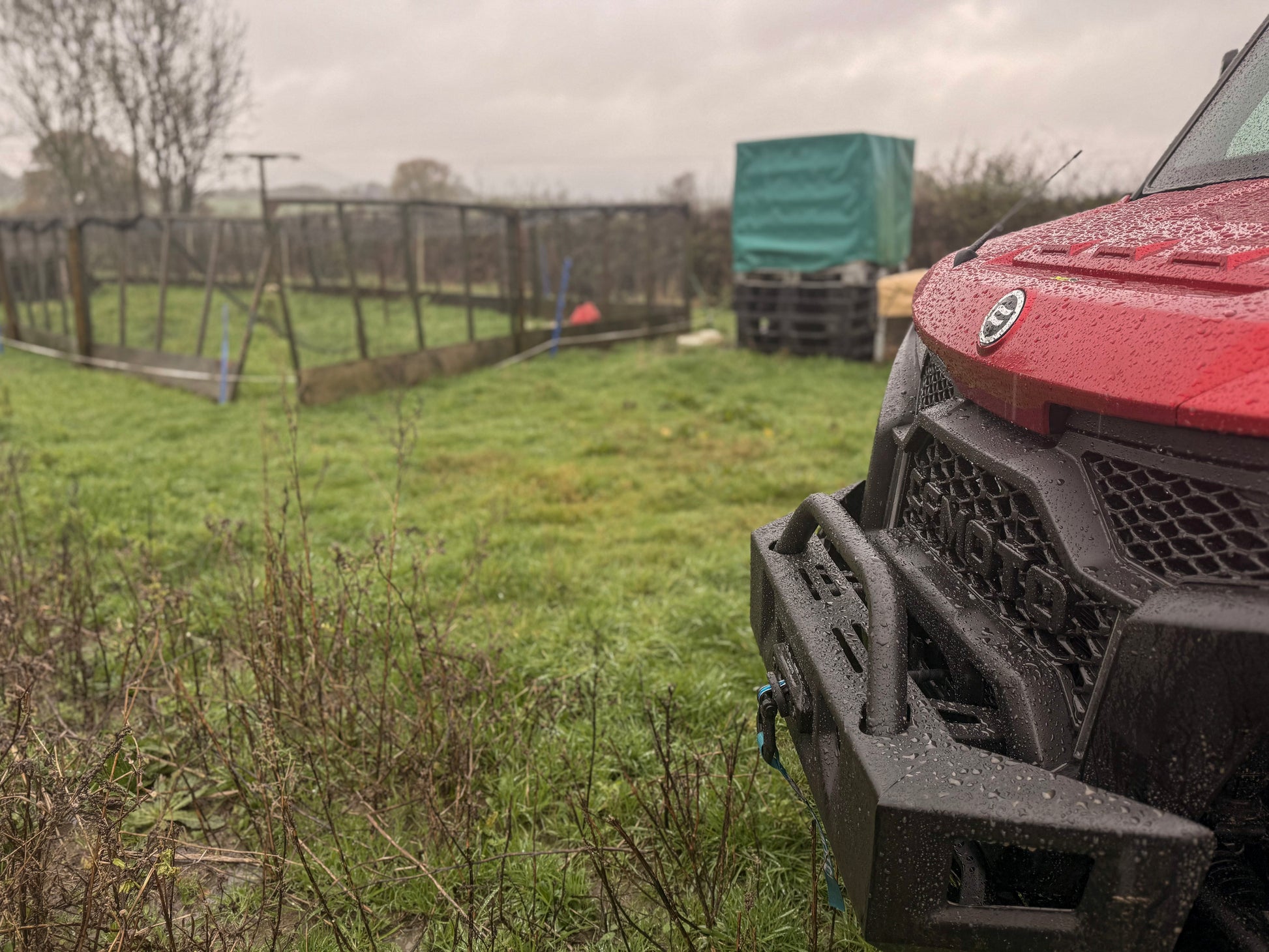 Red CFMOTO off-road vehicle parked on a grassy field with a wooden fence and trees in the background.