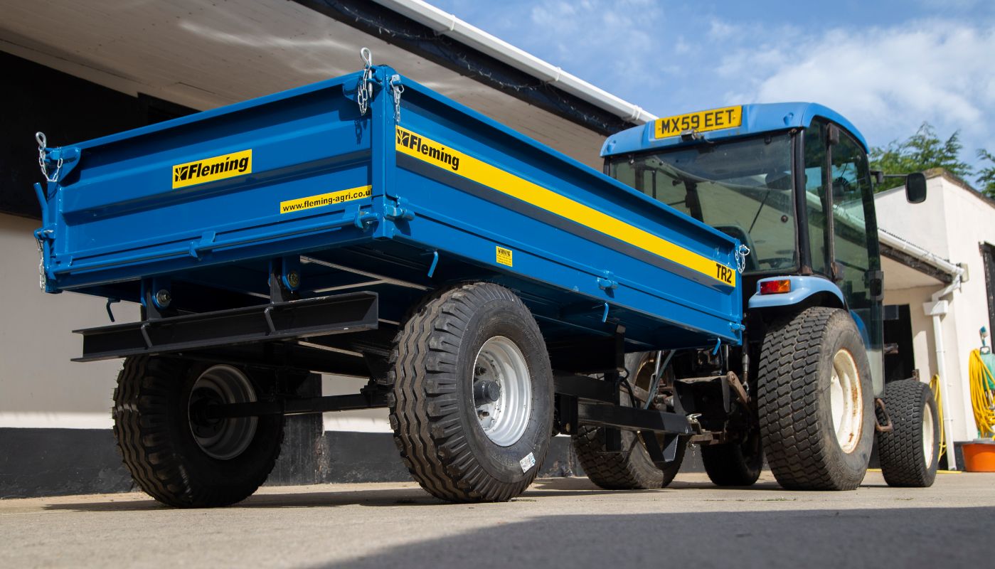 Blue tractor with a Fleming tractor trailer attached, parked under a shelter with a clear sky.