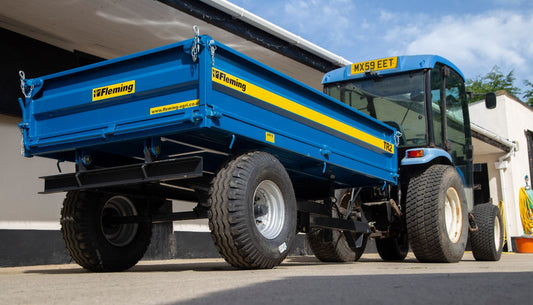 Blue tractor with a Fleming tractor trailer attached, parked under a shelter with a clear sky.