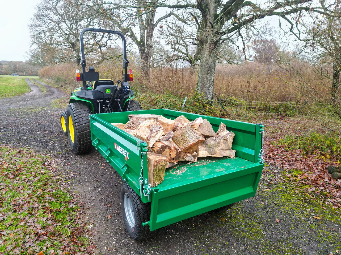 Green tractor with a Wessex tipping tractor trailer carrying wood logs on a rural road.