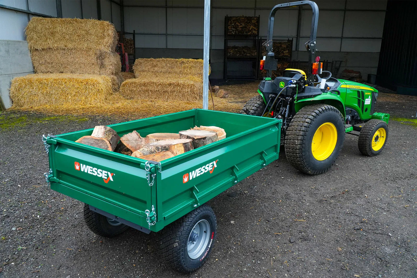Wessex tipping tractor trailer attached to a small tractor with logs inside, in a barn setting.