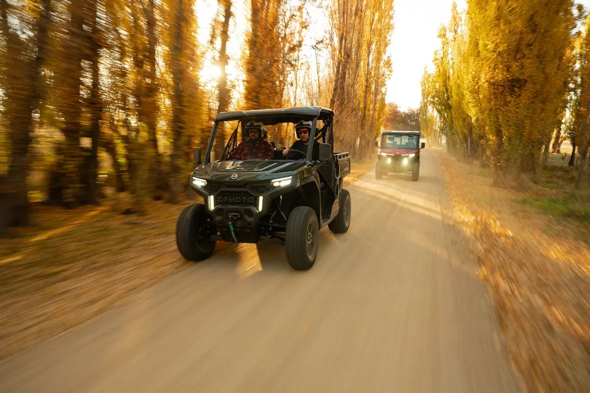 Two CFMOTO off-road vehicles driving on a dirt road lined with trees during sunset.
